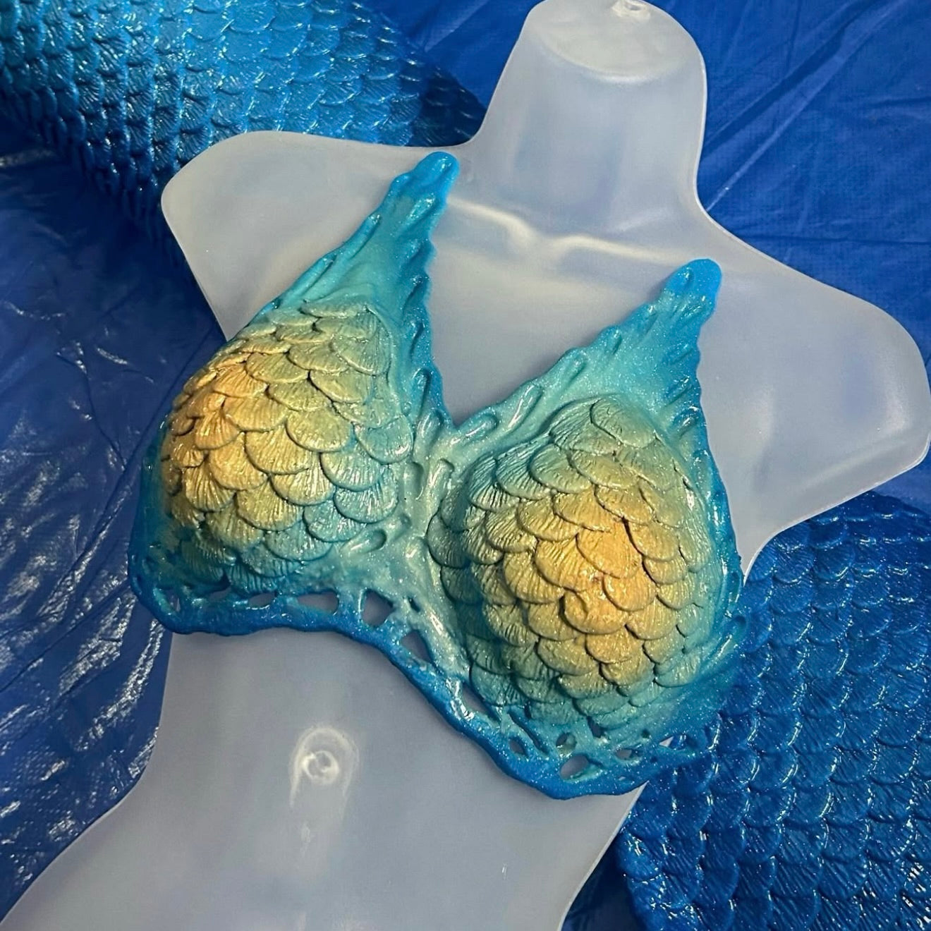 Mermaid scale-patterned bra on a mannequin against a blue, yellow and teal. Custom Mako mermaid cosplay top, realistic silicone design for pool performances